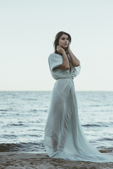 Fototapeta na wymiar Beautiful woman in long white flowing dress standing on the beach