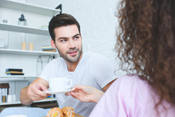 cropped shot of handsome young man holding cup of coffee and looking at girlfriend in  bedroom