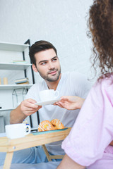 handsome young man holding cup of coffee and looking at girlfriend in  bedroom