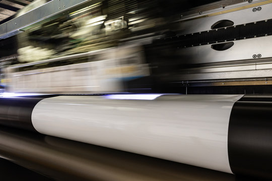 Massive print head flashing by while processing a big oversized roll of glossy premium paper in a professional plot and print facility.