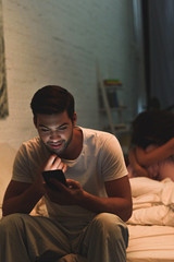 young man using smartphone while upset girlfriend sitting on bed behind, relationship difficulties concept