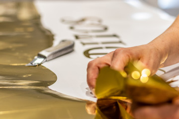 Girl cutting out letters from a golden roll of vinyl. Small hands and cutter on shiny background.