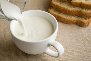 Pouring milk into a large white cup on the background of bread.