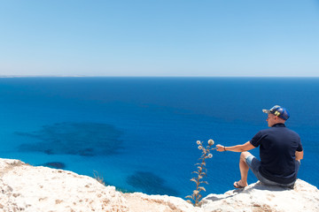 Bald man in a T-shirt and shorts sitting alone on top of a mountain overlooking the sea