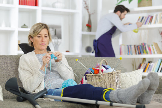 Injured lady knitting while man dusts shelves in background