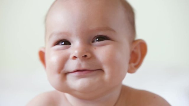 A cute little baby is looking into the camera and is happy on a white bed sheet smiles close up portrait
