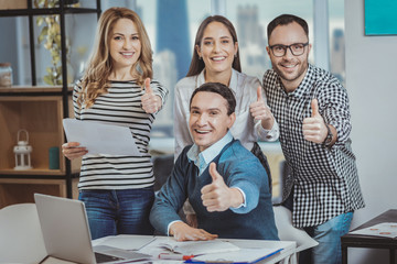 Fototapeta premium Best team. Energetic four colleagues looking at camera and demonstrating thumb