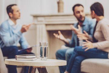 Fresh water. Glass of water standing on the table with unhappy emotional couple arguing in the background