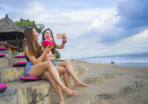 two young happy and attractive Asian Chinese and Korean women hanging out , girlfriends enjoying holidays trip in tropical resort taking selfie picture with mobile phone
