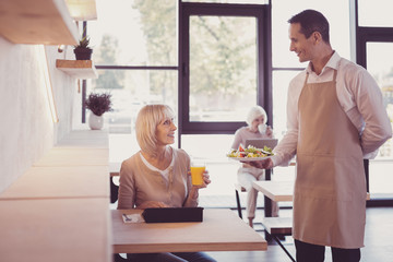 Your order. Responsible handsome pleasant waiter standing in the cafe near a woman who sitting by the table smiling and holding a dish.