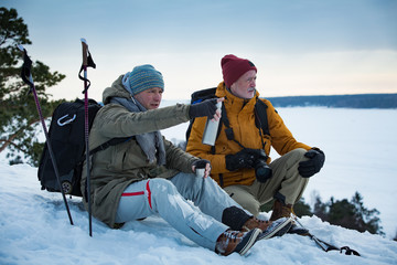 Fototapeta na wymiar Two mature men exploring Finland in winter. Hikers sit on top of rock, take pictures with camera, drink hot coffee from thermos flask. Northern landscape with frozen Baltic Sea and snowy islands.