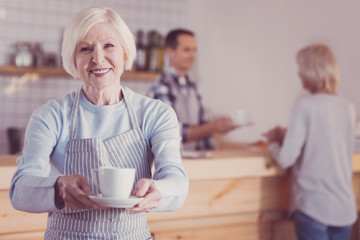 Coffee for you. Joyful friendly aged woman standing in the bright room smiling and holding a cup.
