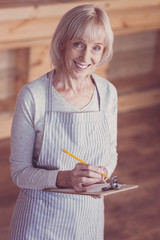 Check everything. Pleasant aged cute woman standing in the empty room smiling and writing.