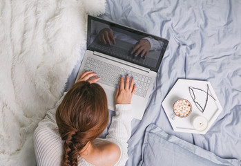 Young woman in white sweater lying on the bed and using laptop.