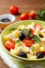 Pasta salad with cherry tomatoes, black olives, feta cheese and basil on wooden background
