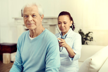 Taking care of health. Selective focus on a serene retired gentleman sitting on a sofa while a female nurse using her stethoscope and listening to his lungs.