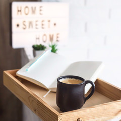 A cup of coffee and an open notebook on a wooden tray in the bright interior of the apartment. Home sweet home written on a decorative frame