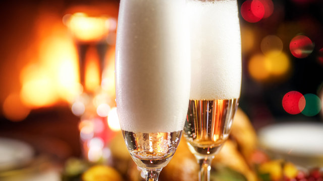 Closeup image of bubbles and foam in glasses of champagne against burning fireplace and Christmas tree