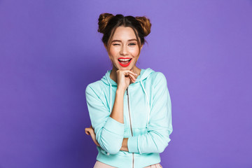 Portrait of happy brunette girl with two buns smiling and winking at you, isolated over violet background in studio