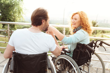 Love you more. Thoughtful mature guy kissing a hand of his smiling wife while both sitting in their wheelchairs and enjoying a pleasant conversation.
