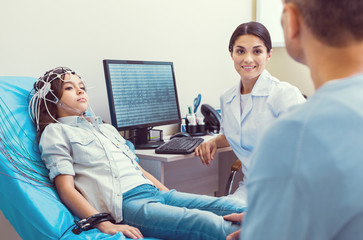 Stressed moment. Smiling female doctor smiling to a father while his poor little girl getting her brain analyzed by an electroencephalograph.