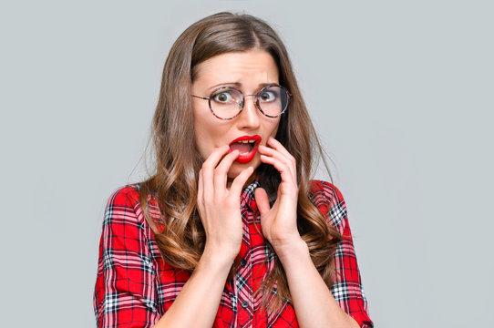 A frightened young woman in round glasses and a red checkered shirt holds her hands near her mouth because of fear