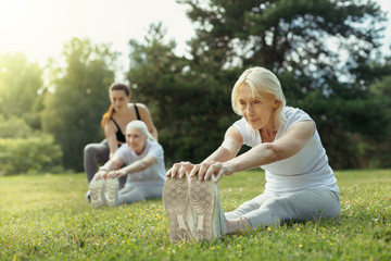 Feeling much better. Concentrated elderly woman sitting on the grass and exercising during a group...