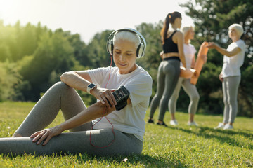 Rhythmical training. Joyful elderly woman sitting on the grass and choosing music before starting her workout in a local park. - Powered by Adobe