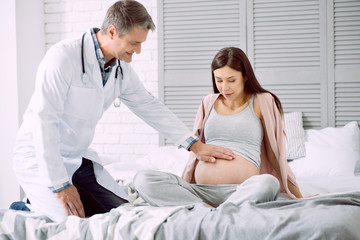 Obraz premium Pregnancy control. Nice positive friendly doctor looking at his patient and putting a hand on her belly while checking her health