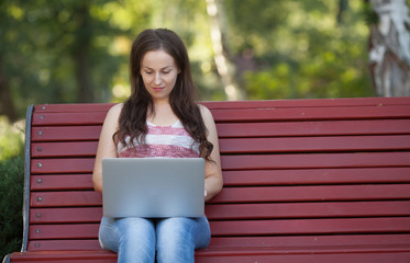 Obraz premium Beautiful caucasian woman is sitting at the bench in a city park with a laptop connected to wi-fi. Entrepreneur is working on a portable computer while sitting outdoors on a blurred urban background.