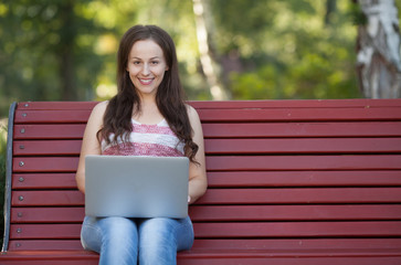 Obraz premium Beautiful caucasian woman is sitting at the bench in a city park with a laptop connected to wi-fi. Entrepreneur is working on a portable computer while sitting outdoors on a blurred urban background.