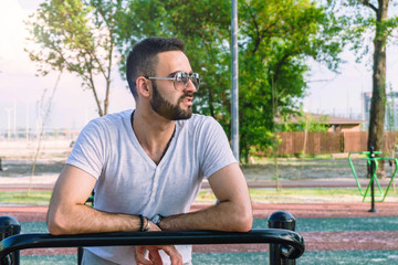 Fototapeta premium a young guy in good physical shape with a beard and in sunglasses trains on the simulators on the street in a city park on a hot sunny day in the summer