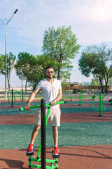 Obraz premium a young guy in good physical shape with a beard and in sunglasses trains on the simulators on the street in a city park on a hot sunny day in the summer