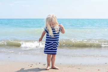 Little blonde girl walking on the beach, holding a seagull's feather