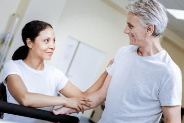 Obraz premium Good results. Emotional active aged man feeling glad while showing good results in a professional gym with his kind friendly trainer standing by his side and touching his right arm