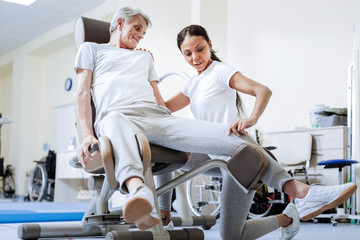Obraz premium Professional help. Calm cheerful experienced doctor in a rehabilitation center touching the knee of her aged disabled patient while helping him to overcome his pain