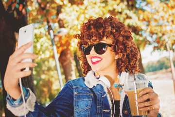 Young curly redhead woman with headphones, smartphone and takeaway coffee, taking a selfie or vlogging. Closeup, vibrant colors, mild retouch.