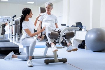 Reliable person. Smiling senior man with injured knee sitting on an exercise machine while a...