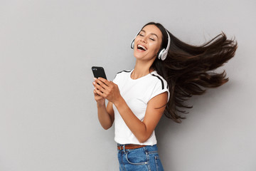 Emotional woman using mobile phone listening music with headphones isolated over grey background.