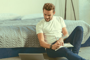 Getting ready for tomorrow class. Relaxed millennial guy sitting on the floor and focusing his attention on a screen of a laptop while writing his home assignment at home.