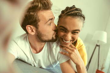 Fototapeta premium Love her so much. Funny young gentleman kissing his radiant soulmate while both relaxing on a bed and posing for a selfie a home.