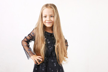 Little blonde girl with long golden hair wearing black dress with stars & planets, dancing, smling & having fun, white wall background. Five years old blonde female child posing. Copy space, close up.