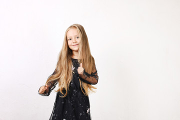 Little blonde girl with long golden hair wearing black dress with stars & planets, dancing, smling & having fun, white wall background. Five years old blonde female child posing. Copy space, close up.