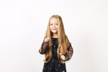 Little blonde girl with long golden hair wearing black dress with stars & planets, dancing, smling & having fun, white wall background. Five years old blonde female child posing. Copy space, close up.