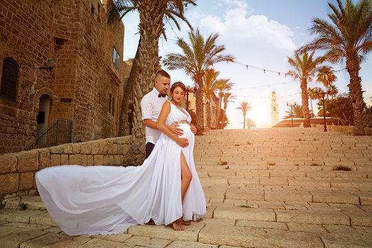 guy hugs a beautiful pregnant wife in a white dress against the backdrop of an ancient city, shchaslivaya pregnant pras on a sunny day