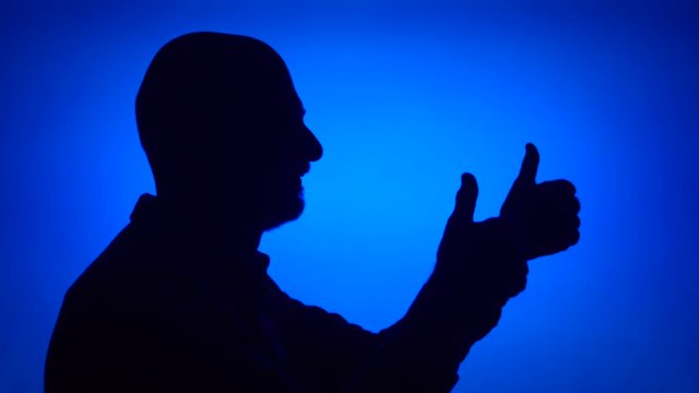 Silhouette of silly senior man having fun making thumb up gestures on blue background. Male's face in profile showing thumbs-up signs. Black contur shadow of grandfather's half-face laughing