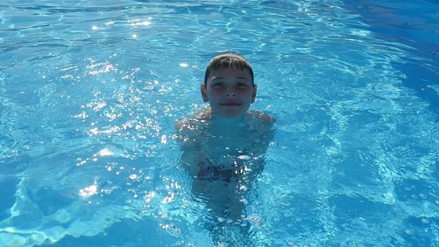 A funny view of a small boy standing and smiling in the celeste waters of a swimming tool sparkling under sunny beams in slow motion