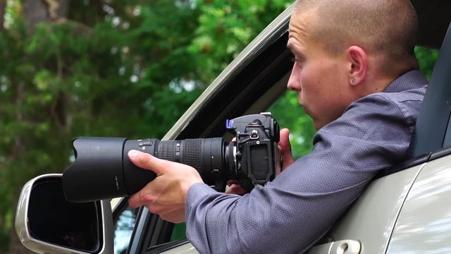 Young Male Spy Sits In The Car Looking Around And Taking Photos To The Camera With A Zoom Lens. Detective Agency Concept