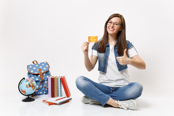 Young pretty smiling woman student in glasses holding credit card showing thumb up sitting near globe backpack, school books isolated on white background. Education in high school university college.