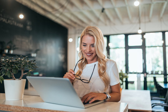 Smiling blonde woman hoding eyeglasses while using laptop.